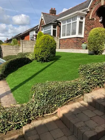 Front garden of a red-brick bungalow with a new artificial lawn above a low wall, framed by clipped shrubs and hedging