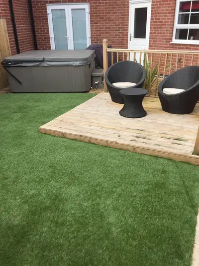 Artificial lawn beside raised timber decking with a hot tub and two rattan chairs, outside a brick house
