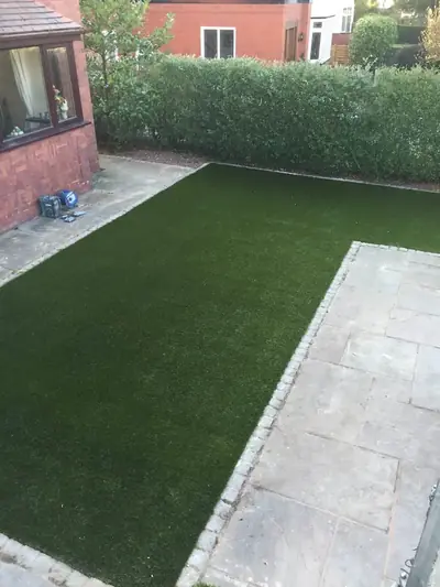 Long artificial lawn beside a wide stone patio, with a tall hedge and a brick house behind