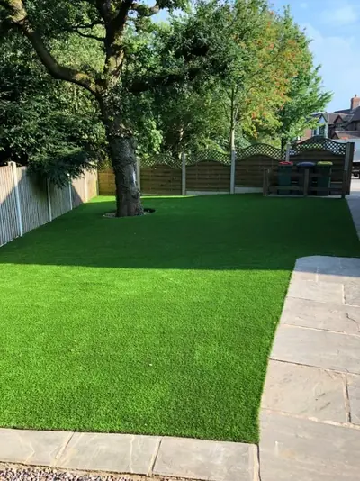 Back garden artificial lawn by Elite Artificial Lawns in summer sun, with a mature tree, a fence and a sandstone patio along one side